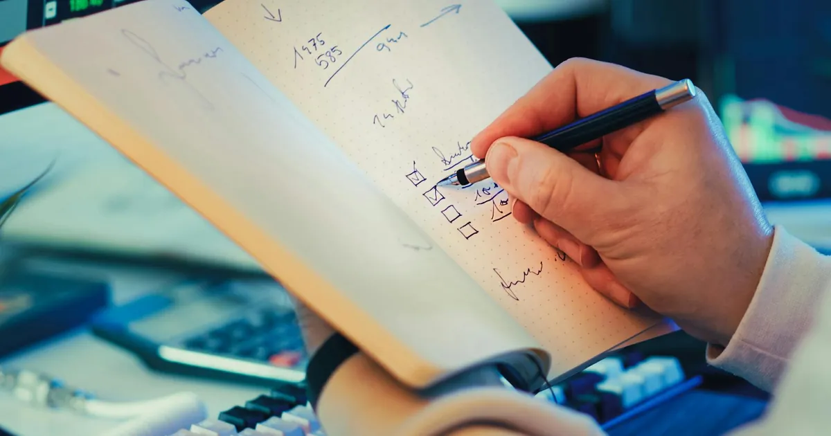 A hand writing in a notebook during stock market analysis on a desk.