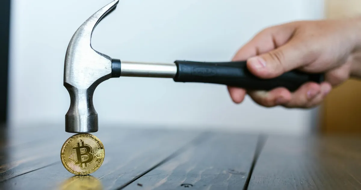 A hand holding a hammer above a Bitcoin coin on a wooden surface, symbolizing cryptocurrency volatility.