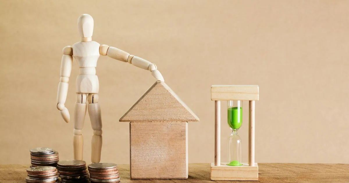 Wooden mannequin with house model, coins, and hourglass symbolizing financial planning.