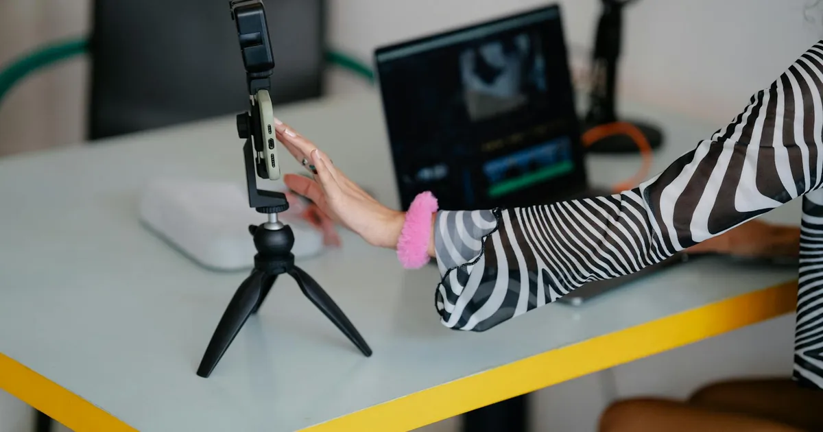 Woman adjusting smartphone on tripod with laptop setup for video editing.
