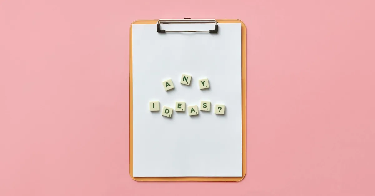 Creative conceptual image of a clipboard with letters spelling 'Any Ideas?' using Scrabble tiles on a pink background.