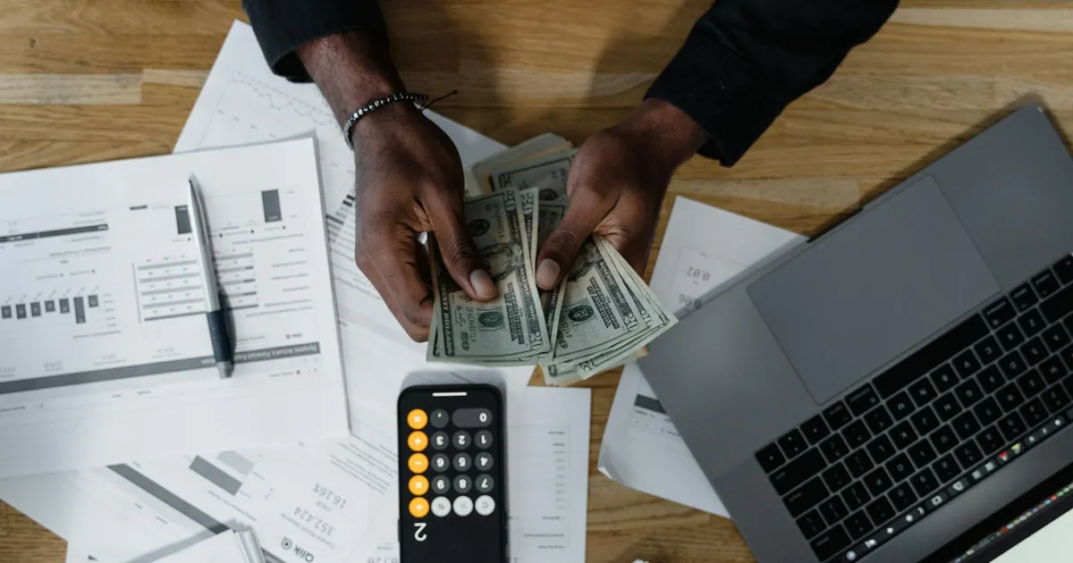 Person counting US dollars, using a calculator and laptop, with financial documents on a wooden desk.